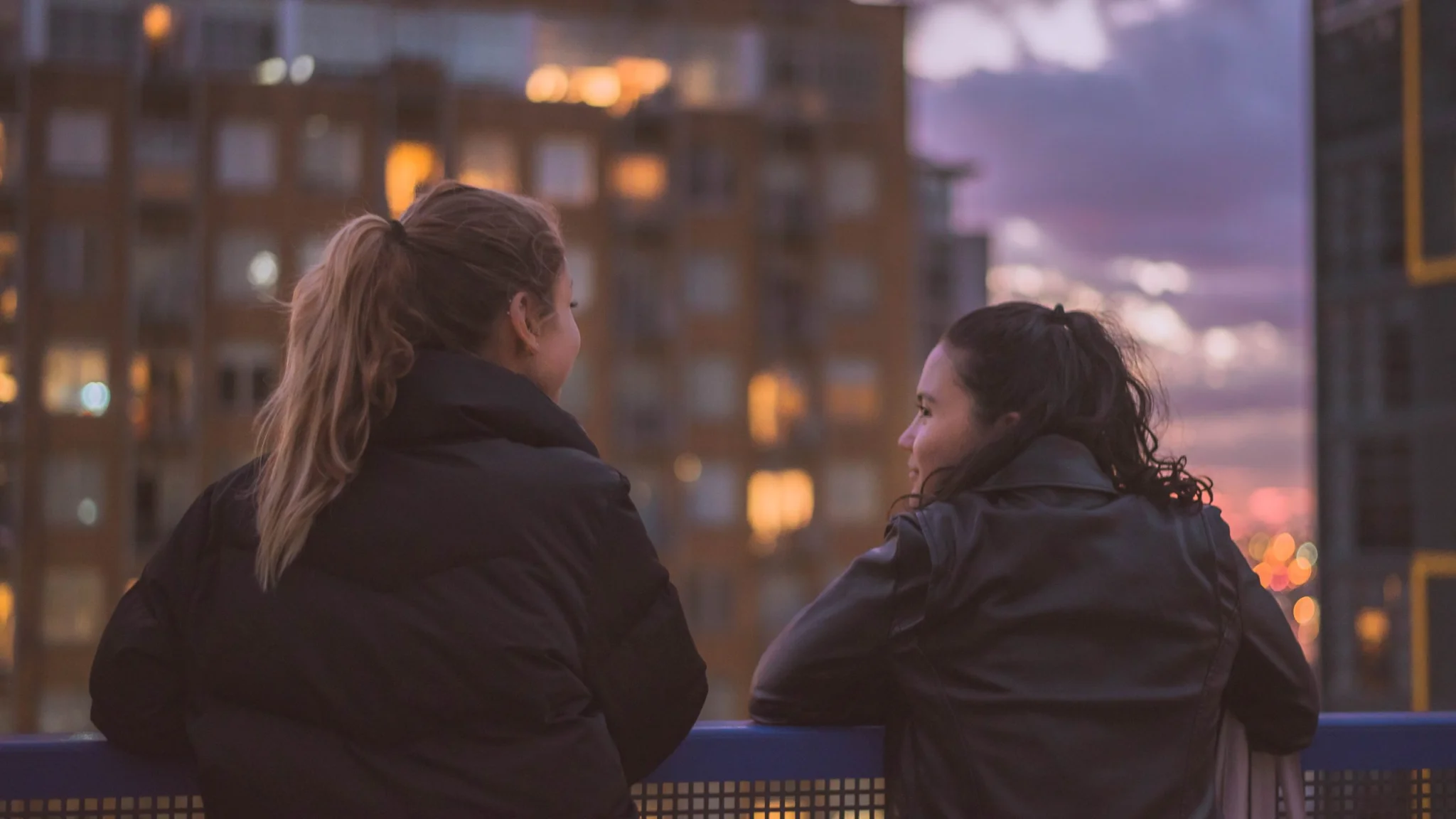 Two young women leaning over a ledge on a rooftop and having a conversation